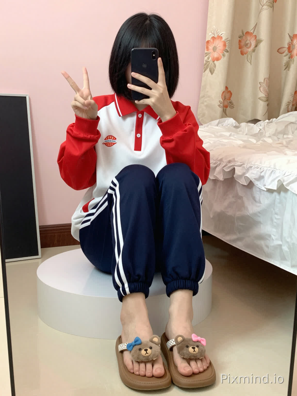 A young woman with short black hair is sitting on a white round platform, taking a self-portrait. She holds a black smartphone in her left hand, which partially covers her face, while her right hand flashes a "V" sign gesture. She is wearing a red and white school uniform jacket, with red cuffs and a red collar, a white middle section, and red buttons on the collar; paired with dark blue sweatpants that have two white stripes running down the legs. Her feet are in brown flip-flops with white polka-dot straps, and the toes of the flip-flops are decorated with plush cartoon elements: two gray bear heads, one with a blue bow and the other with a pink bow.
The background features a light pink wall, a bed with white ruffled sheets on the right side, and beside the bed, a floral-patterned curtain in beige and orange tones. On the left side of the image, there is a black rectangular object (likely a drawing board or mirror). The floor is covered with light-colored tiles, and the overall lighting is warm and soft, creating a cozy daily indoor scene with a realistic style.